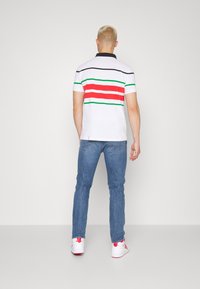 Bílá polo košile s červenými, zelenými a černými pruhy, doplněná modrými džínami. Model má na sobě bílé tenisky s růžovými detaily.