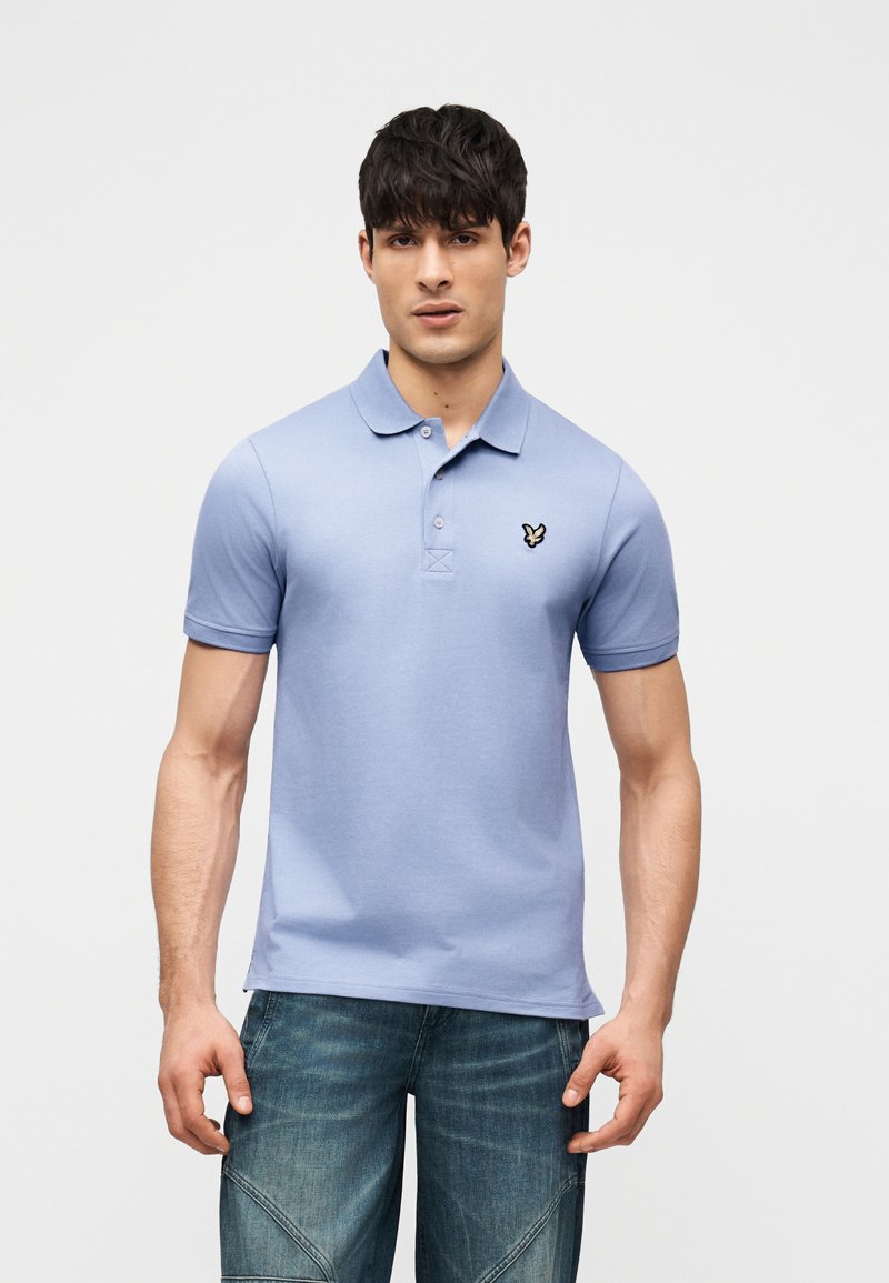 Νεαρός άνδρας που φορά ένα ανοιχτό γαλάζιο κοντομάνικο πόλο με μικρό λογότυπο στο στήθος και μπλε τζιν, σε ουδέτερη στάση.