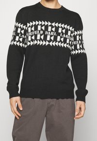 Czarny sweter z dzianiny z białym geometrycznym wzorem na klatce piersiowej i rękawach. Zakończony ściągaczami na mankietach i dolnym brzegu.