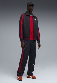 Φόρμα Puma που περιλαμβάνει ένα ναυτικό μπουφάν με κόκκινες ρίγες, συνδυασμένο με ένα κόκκινο μπλουζάκι, ναυτικό παντελόνι και κόκκινα αθλητικά παπούτσια. Μαύρο καπέλο με λογότυπο.