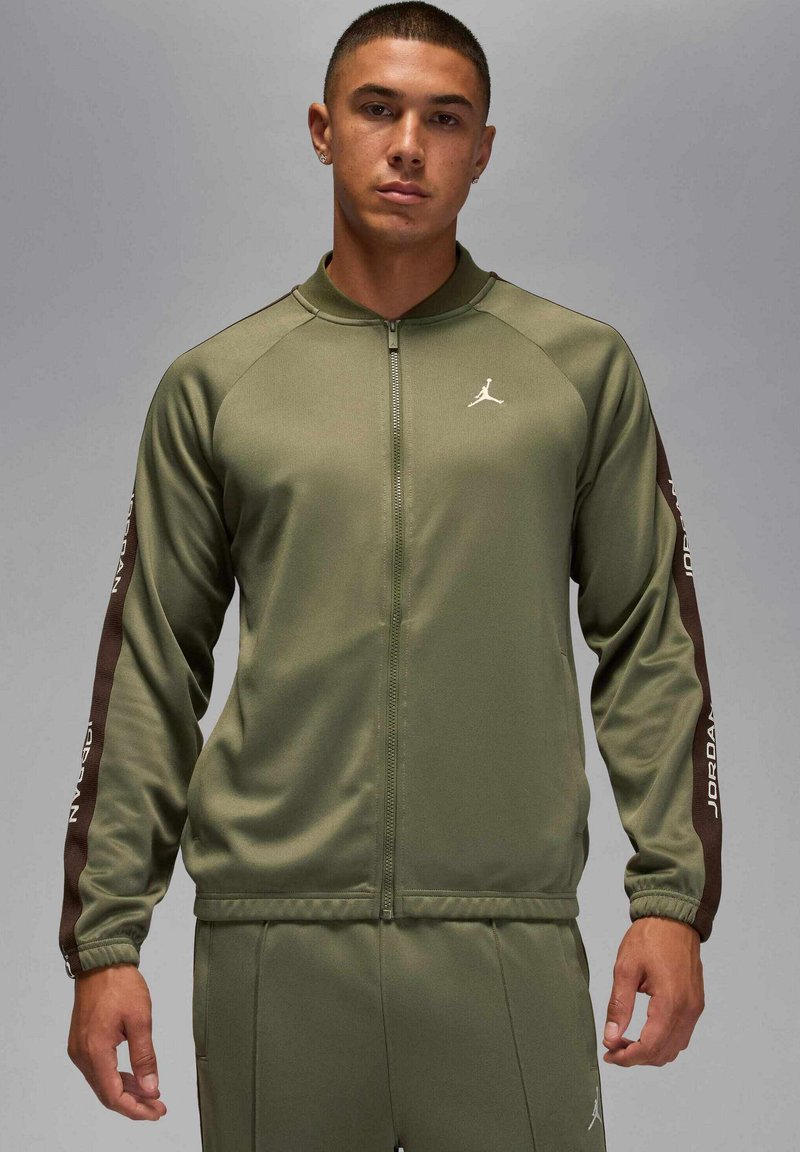Olivgrön zip-up träningsjacka med bruna ränder på sidorna. Har Jordan-märkning på bröstet och ärmarna, ribbade muddar och slät textur.