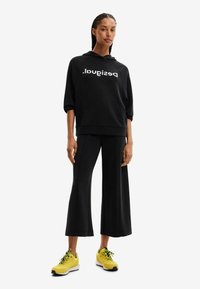 Felpa nera con cappuccio e logo bianco, abbinata a pantaloni larghi neri e scarpe da ginnastica gialle con lacci verdi.