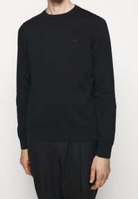 Mann in einem schlichten schwarzen Langarmpullover mit kleinem "HUGO"-Logo auf der Brust und schwarzen Hosen, der vor einem weißen Hintergrund steht.