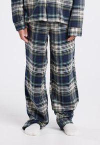 Persona vestida con pantalones de pijama a cuadros en azul, blanco y verde y una camisa a juego, de pie descalza sobre un suelo blanco.