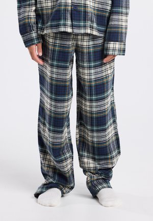 Persoon in blauwe, witte en groene geruite pyjamabroek en bijpassend shirt, staand op blote voeten op een witte vloer.
