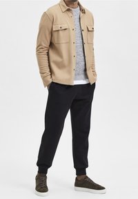 Man som bär en beige knappad skjortjacka, grå stickad tröja, svarta joggerbyxor och bruna sneakers, stående med händerna i fickorna mot en vit bakgrund.