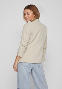 Blazer bej, croit cu guler, mâneci lungi și o siluetă ajustată; asortat cu blugi din denim de culoare albastru deschis.