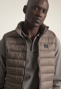 Chaleco acolchado marrón con cierre de cremallera y cuello alto, usado sobre un suéter gris que presenta un logo circular y una tapeta de botones.