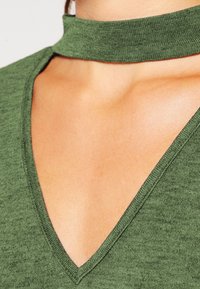 Grüner Strickoberteil mit Choker-Ausschnitt und tiefem V-förmigem Schnitt. Die Textur ist weich mit einem subtilen Melange-Muster.