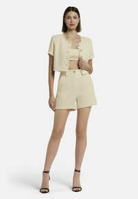 Crèmekleurige cropped jas en bijpassende shorts, gestructureerde stof, knoopsluitingen, gecombineerd met zwarte gespsandalen met hoge hakken.