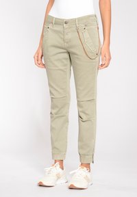 Kaki tapered byxor med framfickor, dragkedjedetalj vid anklarna och en brun kedja som accent. Tillverkad av lättviktig tyg. Stylad med vita sneakers.