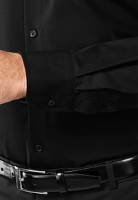 Chemise noire à manches longues avec une texture lisse, dotée d'un poignet à boutons et d'un col à boutons. Portée avec une ceinture noire brillante.
