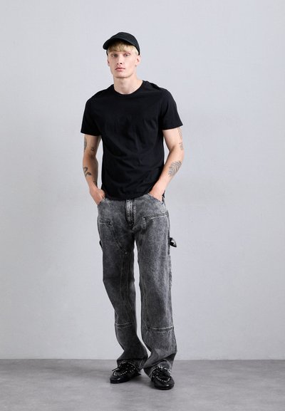 Un jeune homme tatoué portant une casquette noire, un t-shirt noir, un jean gris ample et des chaussures noires à clous, se tenant avec les mains dans les poches contre un mur gris.