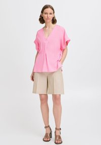 Rosa kortærmet bluse med V-hals og plissédetalje, parret med beige skræddersyede shorts og brune hæle. Ensfarvet baggrund.