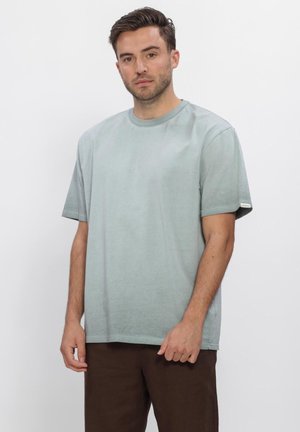 Uomo con capelli corti e scuri che indossa una maglietta grigia chiara oversize e pantaloni marrone scuro, in piedi su uno sfondo bianco uniforme.