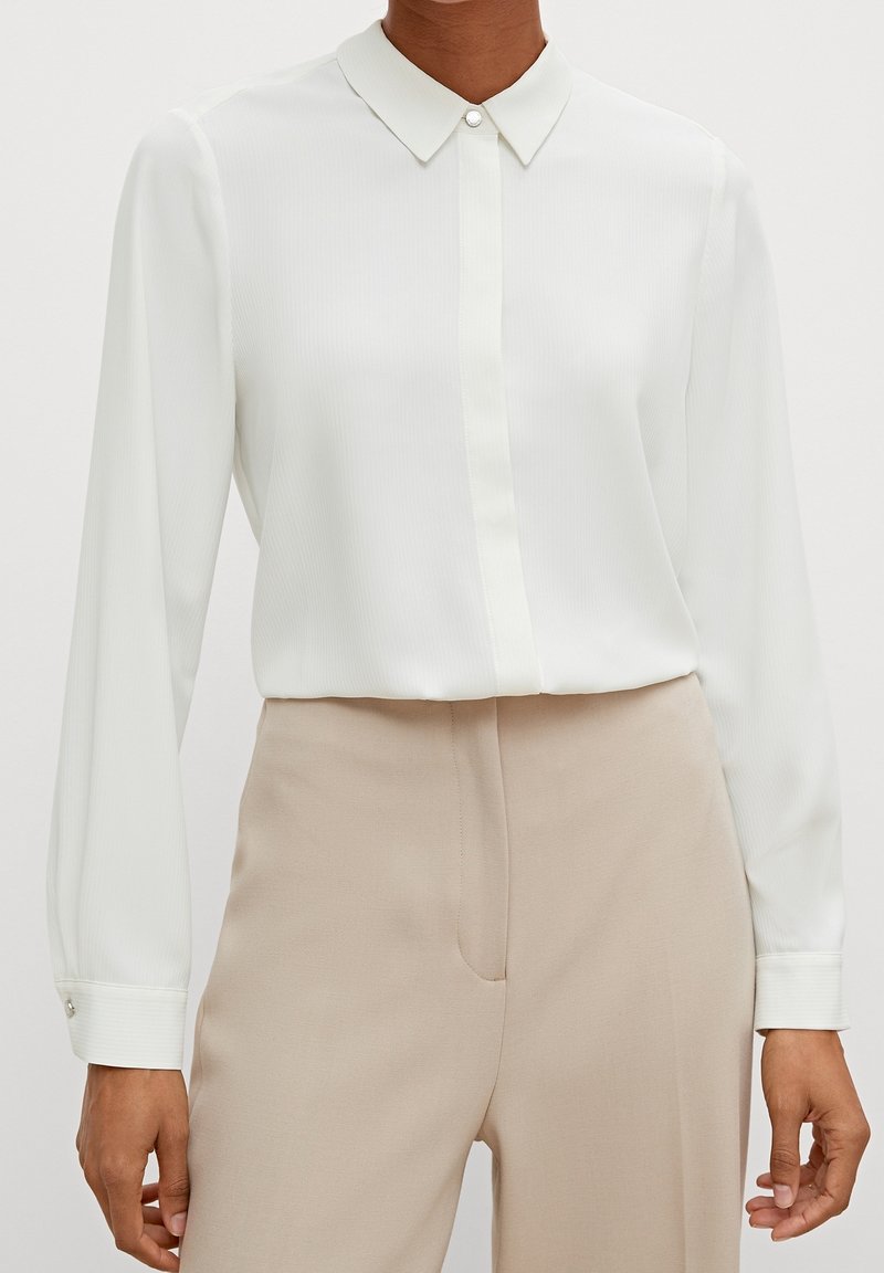 Blouse blanche à manches longues avec un col boutonné, présentant une fine texture côtelée verticale et un petit détail de bouton sur le poignet.
