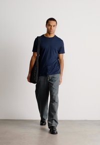 Homem a caminhar para a frente, vestindo uma t-shirt azul-marinho, calças cinzentas, sapatos pretos e a carregar uma mala preta atravessada, contra um fundo claro e neutro.