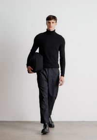 Suéter preto de gola alta, calças pretas com bolsos, botas pretas polidas e uma bolsa preta segurada na mão esquerda. Fundo simples.