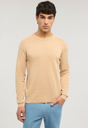 Joven con suéter beige de cuello redondo y jeans azul claro, de pie con una mano en el bolsillo contra un fondo liso y claro.