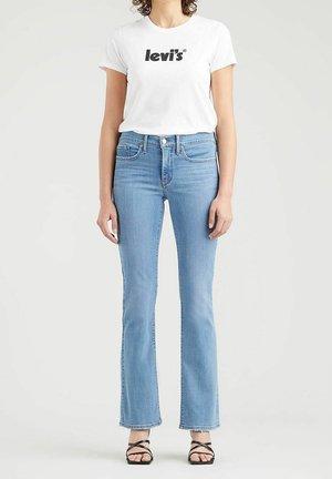 Femme portant un t-shirt blanc Levi's, un jean bleu clair à coupe droite et des sandales noires à brides ouvertes aux orteils, debout devant un fond neutre.