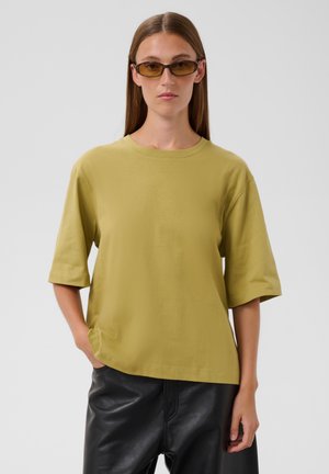 Femme aux cheveux bruns lisses portant de petites lunettes de soleil marron, une chemise ample à manches courtes vert olive et un pantalon en cuir noir, debout avec la main dans la poche.