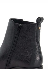 Black leather ankle boot with textured surface, side zipper, and a small gold logo accent. Rounded toe and soft upper edge.