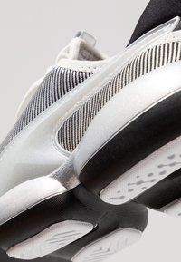 Detailní pohled na podrážku a bok sportovní bílé a stříbrné tenisky s černými a bílými texturovanými látkovými detaily.