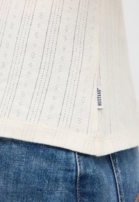 Biały sweter z dzianiny w pionowe paski z małymi otworami, z bocznym rozcięciem i niebieską metką. W połączeniu z niebieskimi dżinsami.