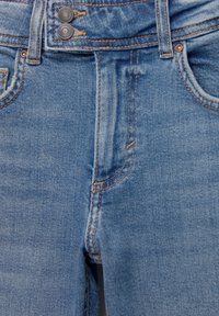 Jean en denim bleu en gros plan montrant une fermeture à double bouton, des poches avant, des passants de ceinture et un détail de couture orange.