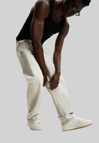Jean en denim de couleur claire avec une coupe décontractée, associés à un débardeur noir et des baskets blanches. Le modèle est présenté dans une pose dynamique.