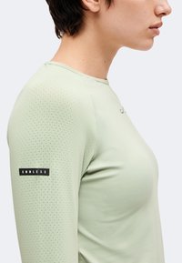 Personne de profil portant une chemise à manches longues vert clair avec de petites perforations sur la manche et une étiquette de texte "ENDLESS" sur le bras.