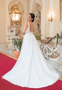 Weißes Brautkleid mit tailliertem Oberteil, Spitzenverzierungen und fließendem Rock. Hat einen tiefen Rücken und zarte Träger. Elegante Umgebung.