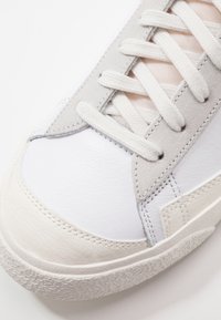 White leather sneaker with light grey suede accents, featuring flat white laces and a textured rubber sole with a patterned grip.