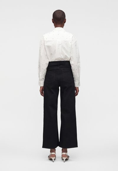 Personne vue de dos portant une chemise blanche à motifs à manches longues, un pantalon noir à jambes larges et des sandales blanches à talons, debout sur un fond uni.