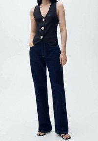 Czarny top bez rękawów z głębokim dekoltem w kształcie litery V i dekoracyjnymi złotymi guzikami, noszony z ciemnoniebieskimi, szerokimi spodniami typu high-waisted.