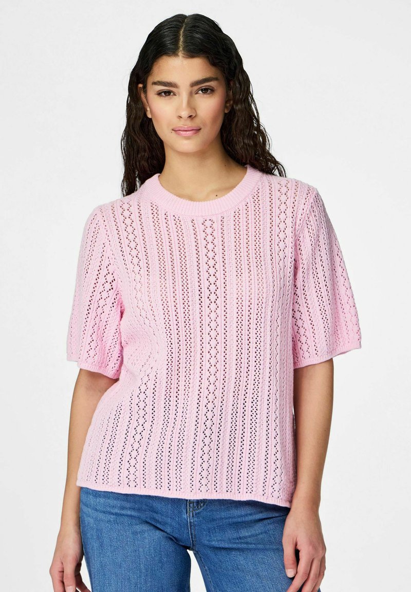 Jeune femme aux longs cheveux foncés portant un haut en maille à manches courtes rose clair avec des motifs en dentelle verticale et un jean bleu, debout devant un fond blanc.