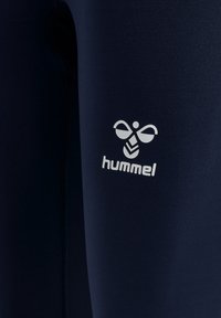 Leggings blu navy realizzati in materiale elastico, con un logo bianco Hummel sulla parte inferiore della gamba e cuciture a contrasto lungo i lati.
