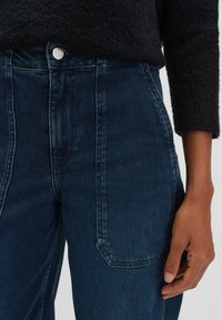 Jeans in denim scuro a vita alta con taglio dritto, caratterizzati da una chiusura con bottone in argento e tasche laterali. La texture appare liscia con leggere sfumature.