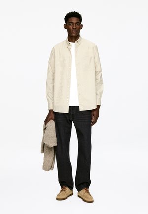 Light beige checkered button-up shirt over a white t-shirt, paired with dark jeans and beige shoes, holding a gray knit sweater.