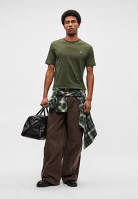 Λαδί πράσινο μπλουζάκι, φαρδιά καφέ παντελόνια, πράσινο καρό πουκάμισο δεμένο στη μέση, μαύρη υφασμάτινη τσάντα, μαύρα παπούτσια. Απλός, χαλαρός σχεδιασμός.