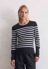 Kvinne iført en svart og hvit horisontalt stripet langermet skjorte og svarte bukser, stående mot en hvit strukturert vegg.