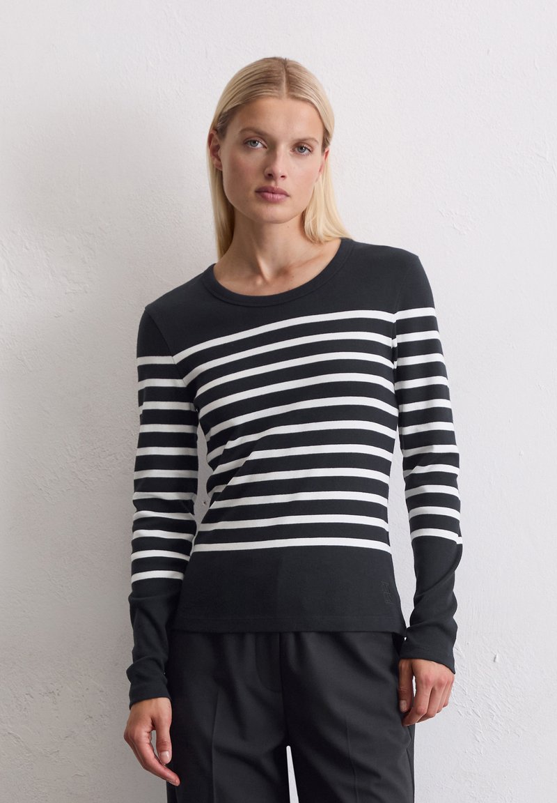 Kvinne iført en svart og hvit horisontalt stripet langermet skjorte og svarte bukser, stående mot en hvit strukturert vegg.