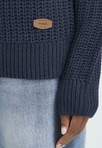 Marineblå strikket sweater med et tekstureret mønster, ribbede ærmer og et læderlogo. Båret over lyseblå denim.