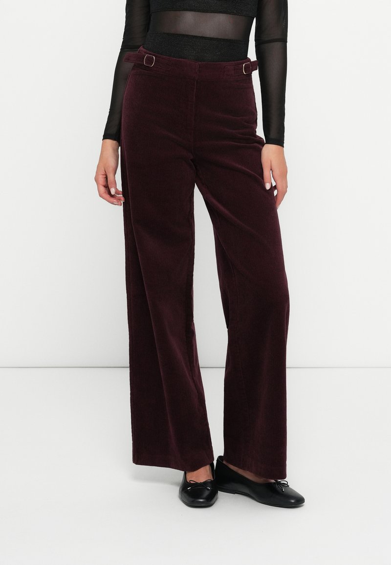 Femeie îmbrăcată cu pantaloni largi din corduroy bordeaux, cu talie înaltă, bluză neagră transparentă cu mâneci lungi și balerini negri, stând în fața unui perete alb.