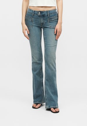 Γυναίκα που φοράει μπλε τζιν καμπάνα με ψηλή μέση και μπροστινές τσέπες και ένα κοντό λευκό τοπ, στέκεται μπροστά σε απλό λευκό φόντο.