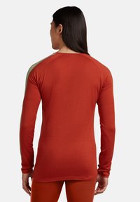 Uomo con capelli lunghi e scuri che indossa una camicia a maniche lunghe arancione bruciato aderente e pantaloni abbinati, visto di spalle su uno sfondo bianco uniforme.