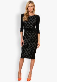 Schwarzes, tailliertes Kleid mit 3/4-Ärmeln, das ein strukturiertes silbernes Muster aufweist. Kombiniert mit orangen, spitzen High Heels, die eine elegante Silhouette betonen.