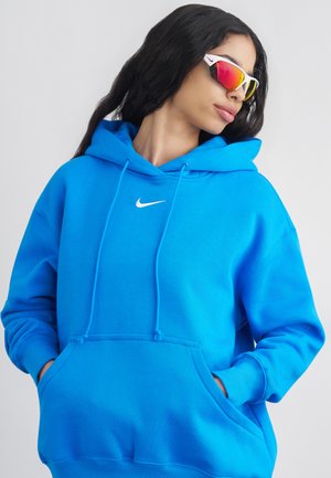 Ženska, oblečena v svetlo modri Nike majici s kapuco in rdečih športnih sončnih očalih z odbojno površino, roke v žepih, gleda v stran.