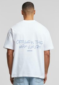 Weißes Baumwoll-T-Shirt mit einem grafischen Textdruck auf der Rückseite in Blau, mit der Aufschrift "OFFLINE IST DER NEUE LUXUS", und locker sitzenden Ärmeln.