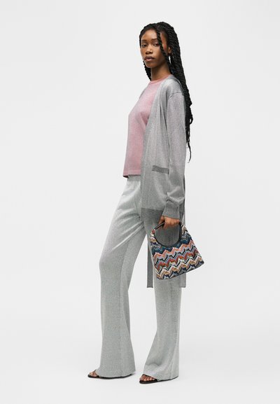 Γυναίκα με μακριά πλεξούδες, ντυμένη με γκρι ζακέτα και παντελόνι, ροζ μπλουζάκι, κρατώντας πολύχρωμη τσάντα με ζιγκ-ζαγκ μοτίβο, όρθια σε λευκό φόντο.